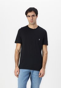 Camiseta de algodón negra con cuello redondo y mangas cortas, que cuenta con un pequeño bolsillo y un sutil detalle de logo en el bolsillo.