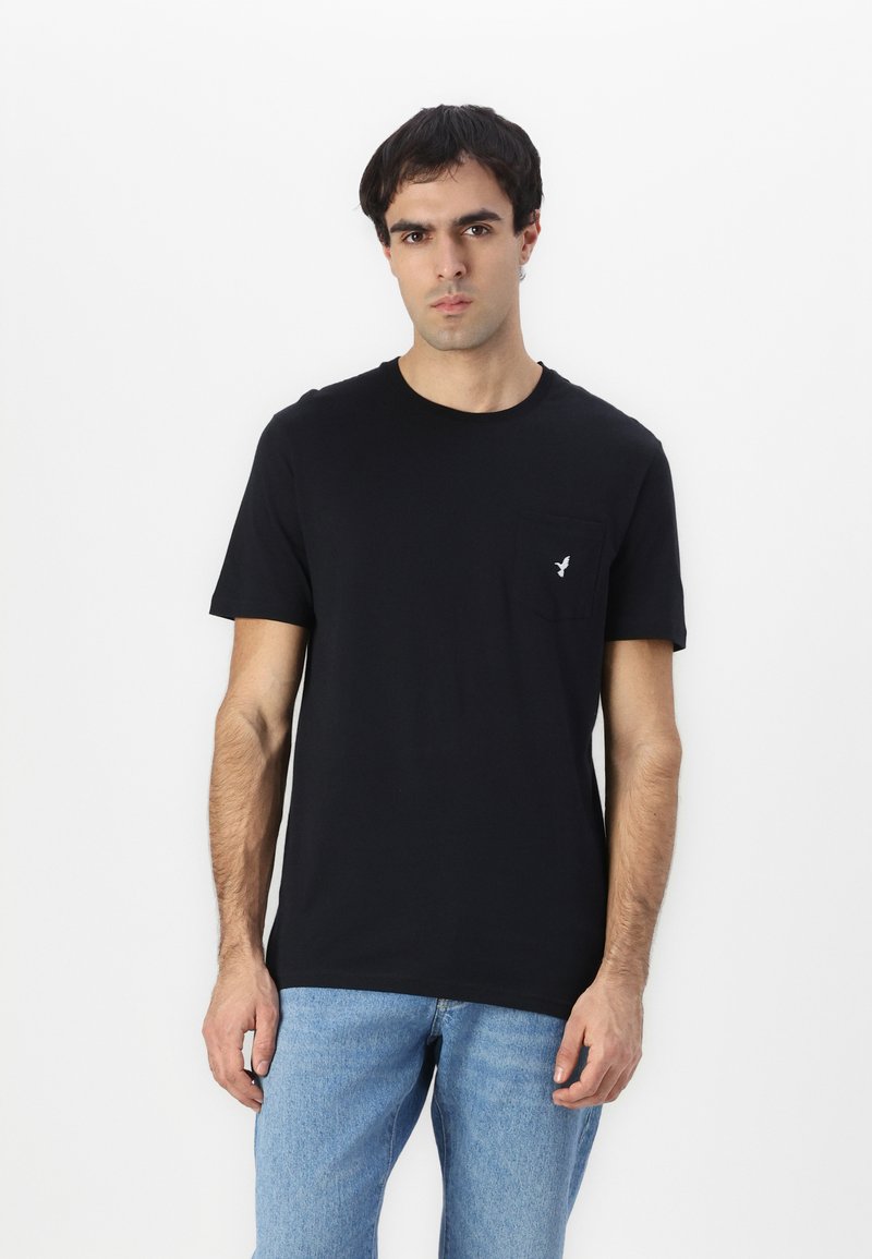 Camiseta de algodón negra con cuello redondo y mangas cortas, que cuenta con un pequeño bolsillo y un sutil detalle de logo en el bolsillo.