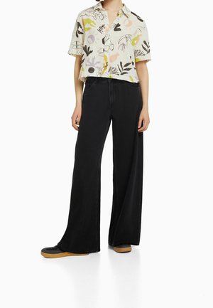 Vrouw draagt een blouse met korte mouwen en abstracte print, gecombineerd met zwarte, hooggetailleerde, wijd uitlopende broek en zwarte sneakers.