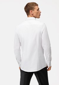 Chemise blanche à manches longues avec un col classique, texture lisse, poignets boutonnés et détail de couture au dos. Associée à un pantalon sombre.