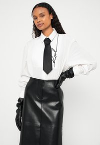 Ženska, oblečena v belo srajco z vezenim napisom "Karl", črno kravato, črno usnjeno krilo in črne usnjene rokavice, pozira z eno roko na boku.