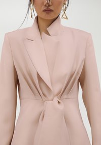 Ružičasti blejzer s čvrstim ramenima, asimetričnom kragnom i detaljem vezanja u struku. Glatka tkanina s čistim linijama i minimalističkim dizajnom.