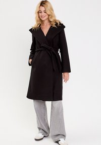 Manteau noir à col large, ceinturé, avec poches, fabriqué en tissu doux ; associé à un pantalon large gris et des baskets.