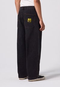 Pantalon en denim noir ample avec de grandes poches arrière, doté d'un patch logo jaune. Le tissu a une texture mate et des coutures solides.