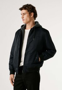 Chaqueta bomber para hombre en azul marino oscuro, con capucha gris desmontable, cierre de cremallera, puños de canalé y detalle sutil del logo en el pecho.