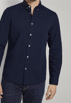 Un homme porte une chemise bleu foncé boutonnée à manches longues avec une poche poitrine, assortie à un jean bleu foncé sur un fond clair uni.