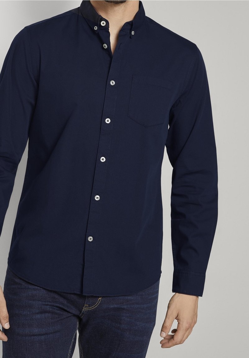 Un homme porte une chemise bleu foncé boutonnée à manches longues avec une poche poitrine, assortie à un jean bleu foncé sur un fond clair uni.