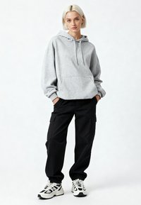 Siva majica s kapuljačom i prednjim džepom, uparena s crnim cargo hlačama s bočnim džepovima. Bijele i crne tenisice upotpunjuju izgled.