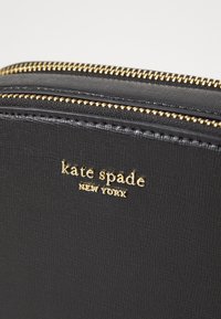 Portafoglio in pelle nera con texture, caratterizzato da una zip dorata, un logo dorato che recita "kate spade NEW YORK" e un design orizzontale.