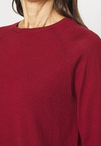 Kobieta ubrana w prostą, długą, rękawową sweter z okrągłym dekoltem w głębokim czerwonym kolorze, z widocznymi szwami wzdłuż ramion.