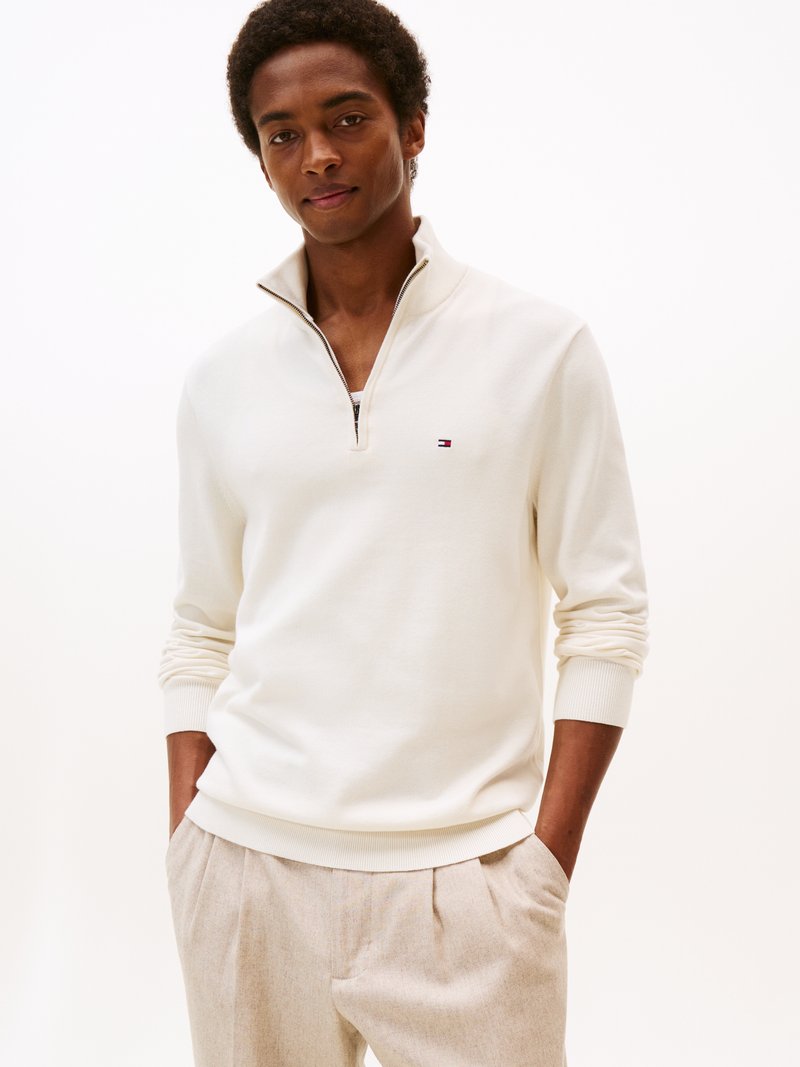 Maglione bianco a zip con collo a 1/4, con polsini e orlo a coste. Pantaloni grigi con pinces. Piccolo dettaglio del logo sul lato sinistro del petto. Texture liscia.