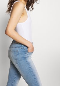 Jeans skinny de lavado claro con una altura media, que presentan una textura suave, diseño de cinco bolsillos y un ligero desvanecimiento. Combinados con una camiseta sin mangas blanca.