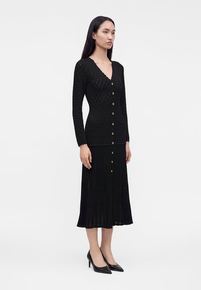 Femme portant un long cardigan noir boutonné avec une taille cintrée et une jupe plissée assortie, associée à des chaussures noires à talons hauts.
