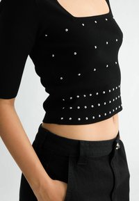 Schwarzes kurzes Oberteil mit quadratischem Ausschnitt, verziert mit silbernen Nieten in einem verstreuten Muster, ergänzt durch hochtaillierte schwarze Hosen.