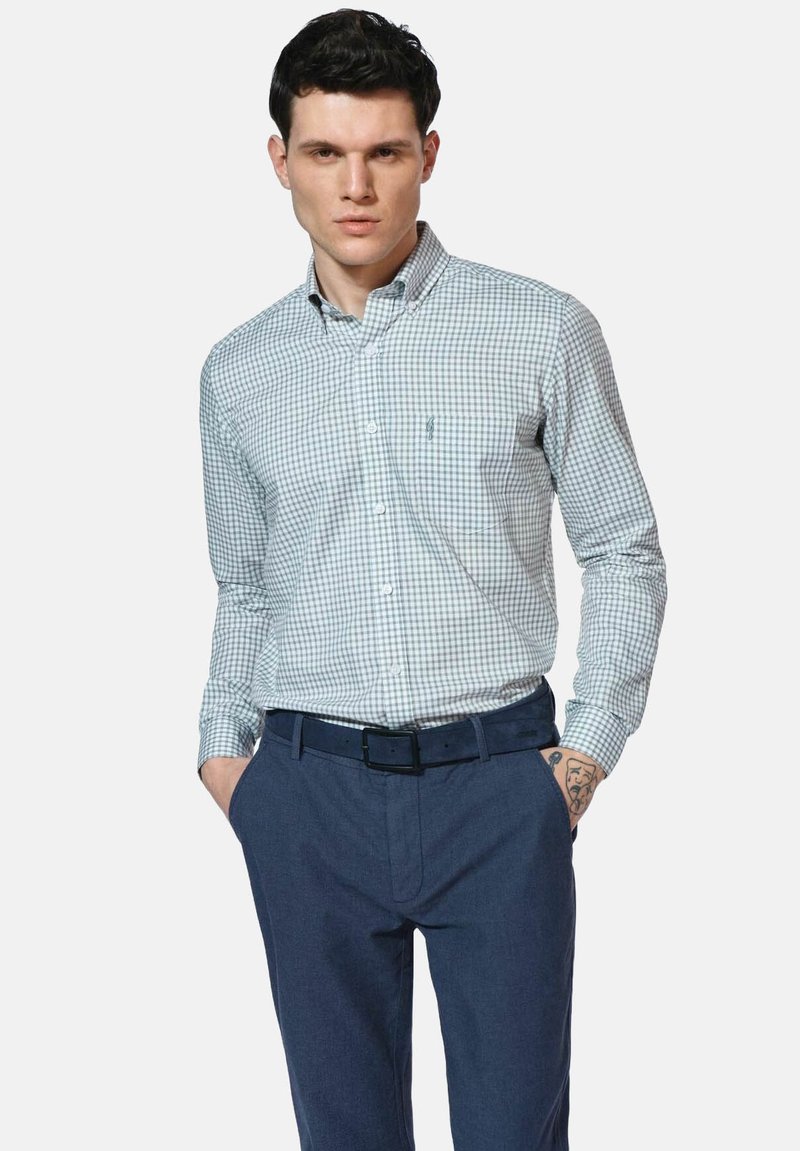 Koszula w kratę z guzikami w zielono-białym kolorze, z długim rękawem. Dopasowany krój z małym logo na klatce piersiowej. Noszona z niebieskimi spodniami i czarnym paskiem.