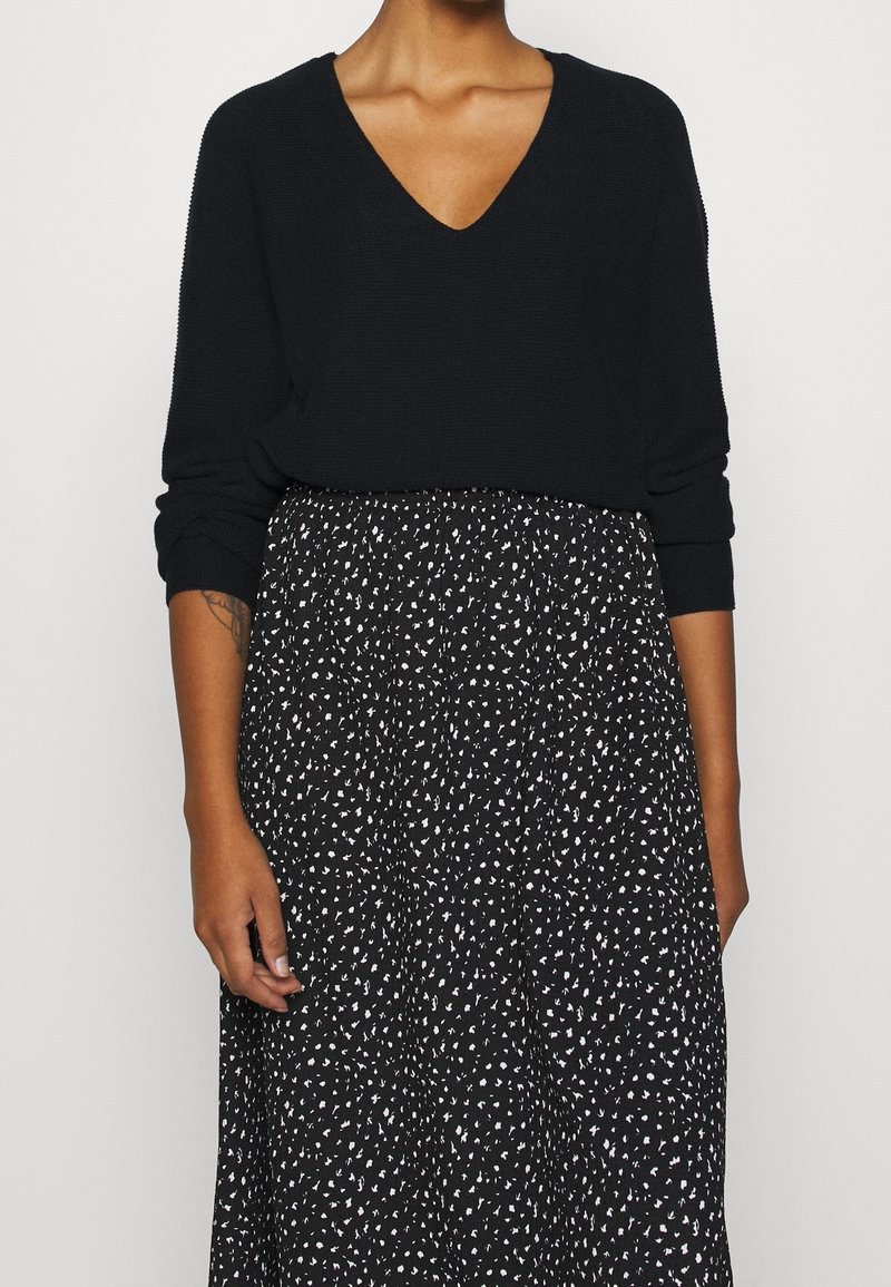 Pull en maille noir avec un col en V et des manches longues, associé à une jupe noire aux motifs abstraits blancs. Texture douce, coupe décontractée.