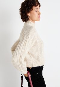 Pulover crem cu model împletit în formă de cablu, cu guler înalt și mâneci lărguțe, asortat cu pantaloni negri din catifea cord și o geantă burgundă.