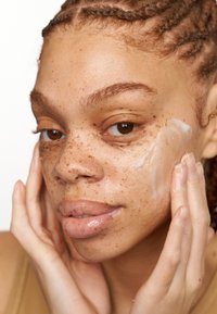 Crème hydratante appliquée sur un visage avec des taches de rousseur ; cheveux bouclés tirés en arrière ; mains touchant doucement la peau, mettant en valeur la texture lisse et humide.