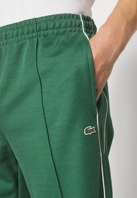 Green athletic joggers in soft material, featuring a side stripe and a small embroidered logo on the thigh. Elastic waistband with drawstring.