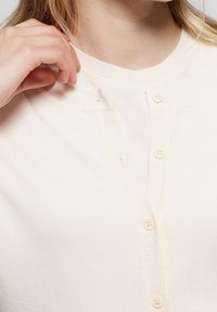 Krémový cardigan z měkkého pleteného materiálu. Má kulatý výstřih, zapínání na knoflíky a jemnou strukturu. Viditelné čtyři knoflíky.