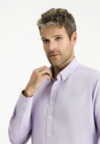 Jasnofioletowa koszula z guzikami, wykonana z fakturowanej tkaniny, z kołnierzykiem o spiczastym kształcie i długimi rękawami. Białe guziki zdobią przednią listwę.