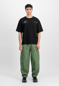 Fekete pamut póló laza szabással, rövid ujjakkal és logónyomattal. Zöld katonai nadrágokkal viselik, amelyek zsebekkel rendelkeznek és laza dizájnnal bírnak.