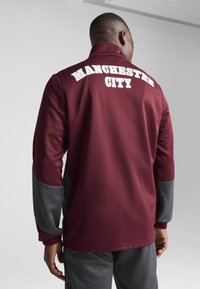 Bordeaux pullover med grå ærmer, har "MANCHESTER CITY" i hvid på bagsiden. Stoffet er glat med en afslappet pasform og høj hals.