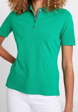 Kobieta ubrana w zieloną koszulkę polo z krótkim rękawem i trzema guzikami oraz białe spodnie, stoi z jedną ręką na biodrze.