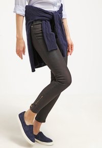 I jeans skinny neri presentano una leggera lucentezza, con cerniere sulle caviglie. Un maglione di maglia blu navy è annodato in vita.