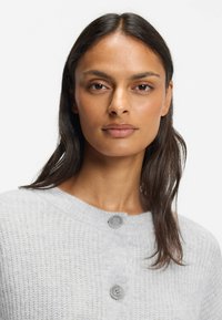 Vaaleanharmaa napillinen neuletakki, jossa on teksturoitu neulottu kangas, pyöreä pääntie ja kaksi näkyvää harmaata nappia rinnan läheisyydessä.