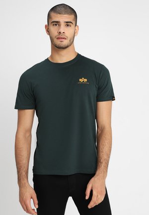 T-shirt in cotone verde scuro a girocollo con maniche corte, con un logo giallo sul lato sinistro del petto e un'etichetta sulla manica sinistra.