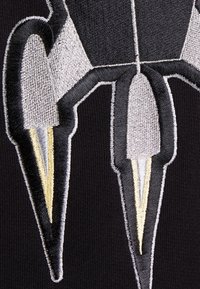 Černá látka s výrazným vyšitým motivem rakety ve stříbrné, šedé a žluté barvě, s detailními texturami a ostrými liniemi.