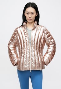 Rózsaarany színű steppelt puffadt kabát fényes kivitelben, magas gallérral és elöl cipzárral. Szürke felső fölött viselve, kék leggingsszel kombinálva.