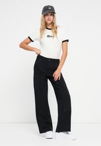 Bela majica s črnim robom, ki nosi logotip "Dickies", v kombinaciji s širokimi črnimi cargo hlačami in belimi športnimi copati, dopolnjena s sivo kapo.