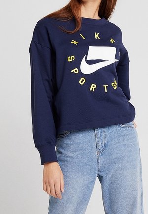 Personne vêtue d'un sweat-shirt Nike Sports bleu marine avec des lettres jaunes et un jean bleu clair, debout devant un fond clair uni.