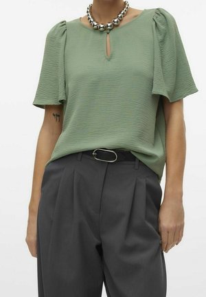 Woman wearing light green textured short-sleeve blouse with keyhole neckline, silver bead necklace, black pleated trousers, and black belt.