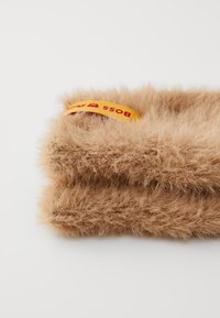 Cachecol de tricot felpudo de cor castanha claro, dobrado, com uma etiqueta de tecido "BOSS" em laranja e vermelho sobre um fundo branco.