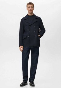 Abrigo marina cruzado de lana con solapas de muesca, bolsillos laterales; combinado con pantalones vaqueros oscuros y zapatos de cuero negros.