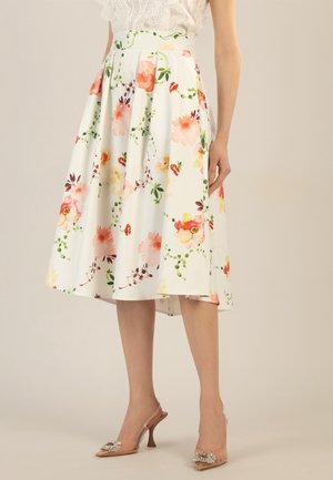 Femme portant une jupe midi blanche à fleurs roses, orange et jaunes, associée à des chaussures à talons beiges avec des détails décoratifs.