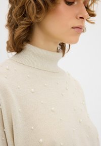 Beige, gebreide coltrui met een textuurstof en verspreide parelachtige versieringen over het oppervlak.