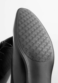 Suela de zapato negra con un patrón texturizado de pequeñas conchas, bordes lisos y una ligera curva. La parte superior muestra material negro y cordones.
