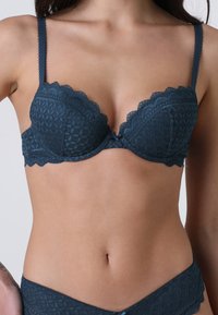 Donna che indossa un reggiseno in pizzo blu scuro e mutande coordinate, mostrando il torso dalle spalle ai fianchi su uno sfondo semplice.