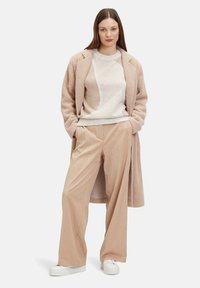 Beiger langer Mantel aus weichem Material über einem gemusterten leichten Strickpullover, kombiniert mit weit geschnittenen beigen Cordhosen und weißen Sneakern.