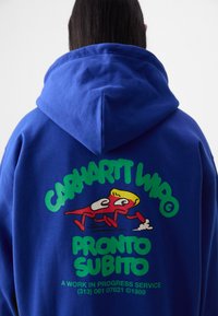 Blå hoodie med en stor grön och röd grafik på baksidan som föreställer en tecknad figur och texten "CARHARTT WIP" och "PRONTO SUBITO."