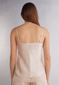 Satinowy top na ramiączkach w delikatnym beżu z regulowanymi ramiączkami i subtelnym koronkowym wykończeniem na dole, prezentujący gładką fakturę i elegancki design.