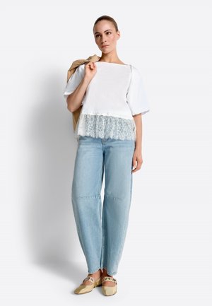 Femme debout portant un haut blanc à manches courtes avec un ourlet en dentelle, un jean bleu clair à jambes larges, des ballerines beiges, tenant une veste beige sur l'épaule.