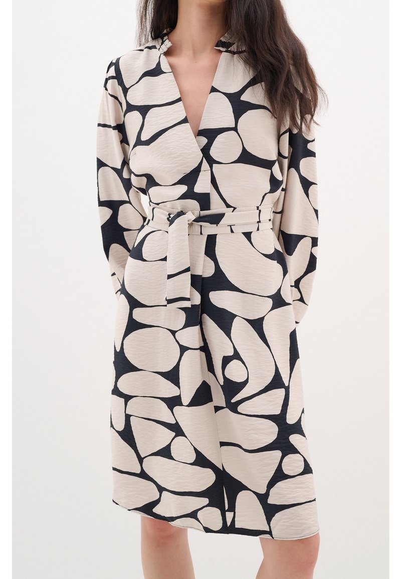 Femme portant une robe portefeuille beige avec des formes abstraites noires irrégulières et une ceinture assortie nouée à la taille, se tenant devant un fond blanc.