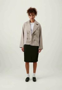 Veste double boutonnage beige clair avec revers larges, associée à une jupe vert foncé arrivant au genou. Le modèle porte des chaussettes blanches et des chaussures noires.