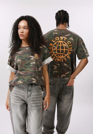 Nő előlről nézve és férfi hátulról látható, mindketten terepmintás pólót viselnek narancssárga földgömbbel és szöveges mintával, szürkés, koptatott farmernadrággal párosítva.