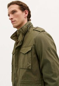 Olijfgroene militaire jas in stijl met een hoge kraag, twee borstzakken, schouderepauletten en een schedellogo op de mouw.